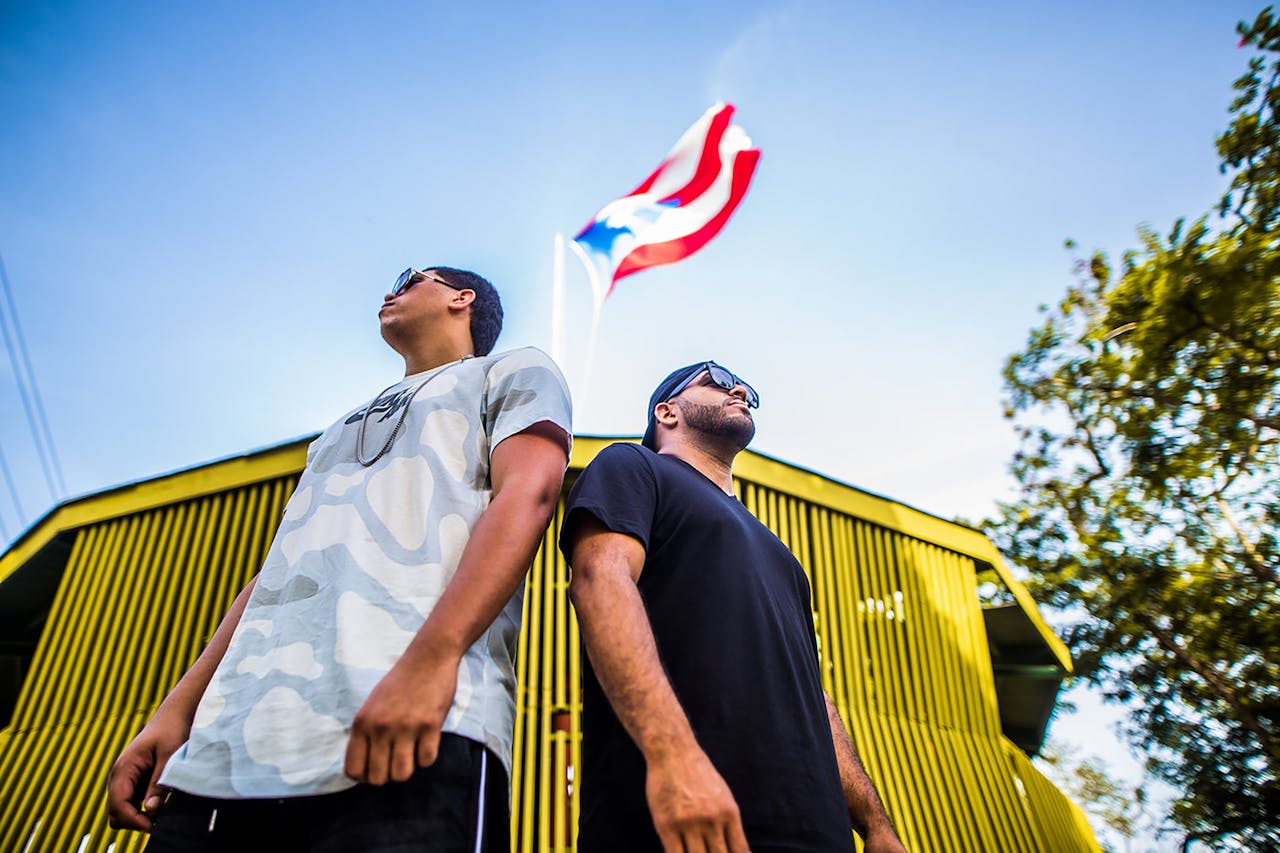 Two men in casual wear pose under the Puerto Rican flag, symbolizing pride and unity.